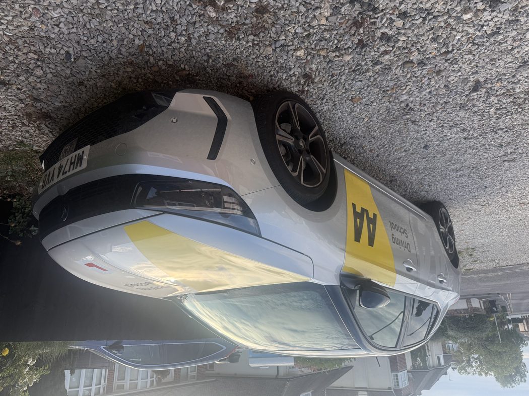 Silver AA Driving School car with yellow livery and L-plate, parked outside a house.